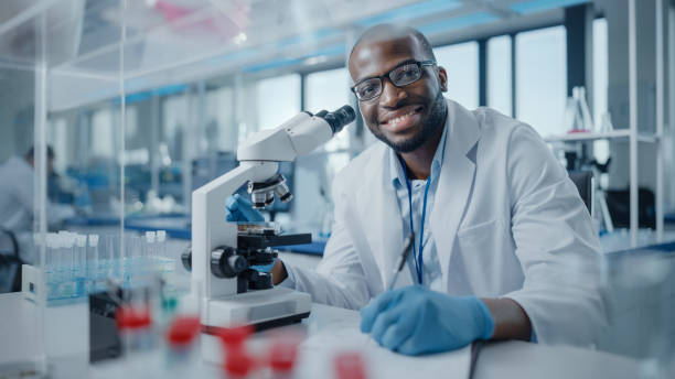Man in witte laboratoriumjas en blauwe handschoenen staat bij microscoop en reageerbuizen in helder laboratorium.