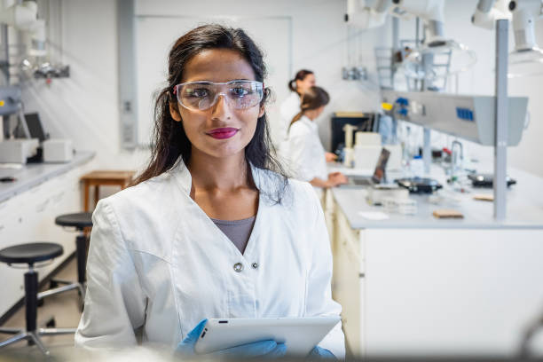 Vrouw in witte labjas en bril met tablet in modern laboratorium met collega’s op de achtergrond