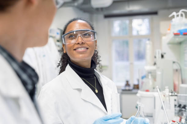 Vrouw in witte labjas met paarse trui en veiligheidsbril werkt in laboratorium met labfles