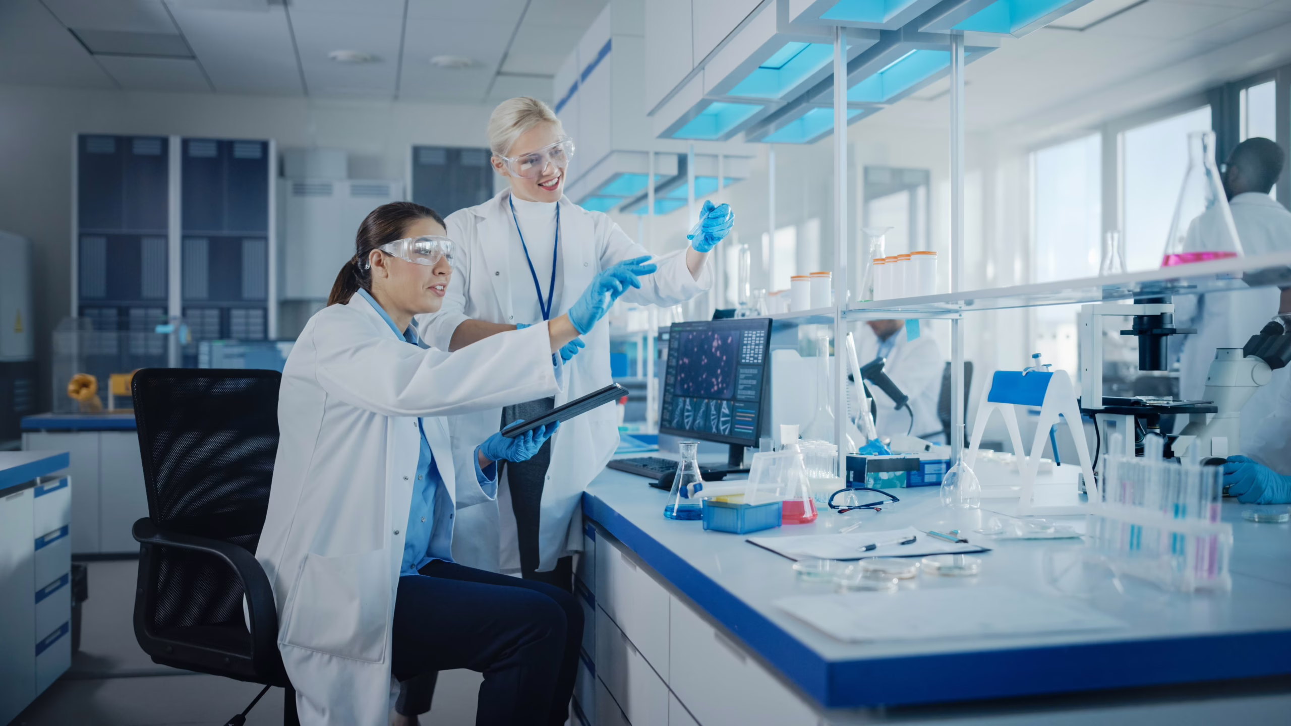 Twee vrouwen in laboratoriumjassen werken samen met pipet, tablet en laboratoriumapparatuur.