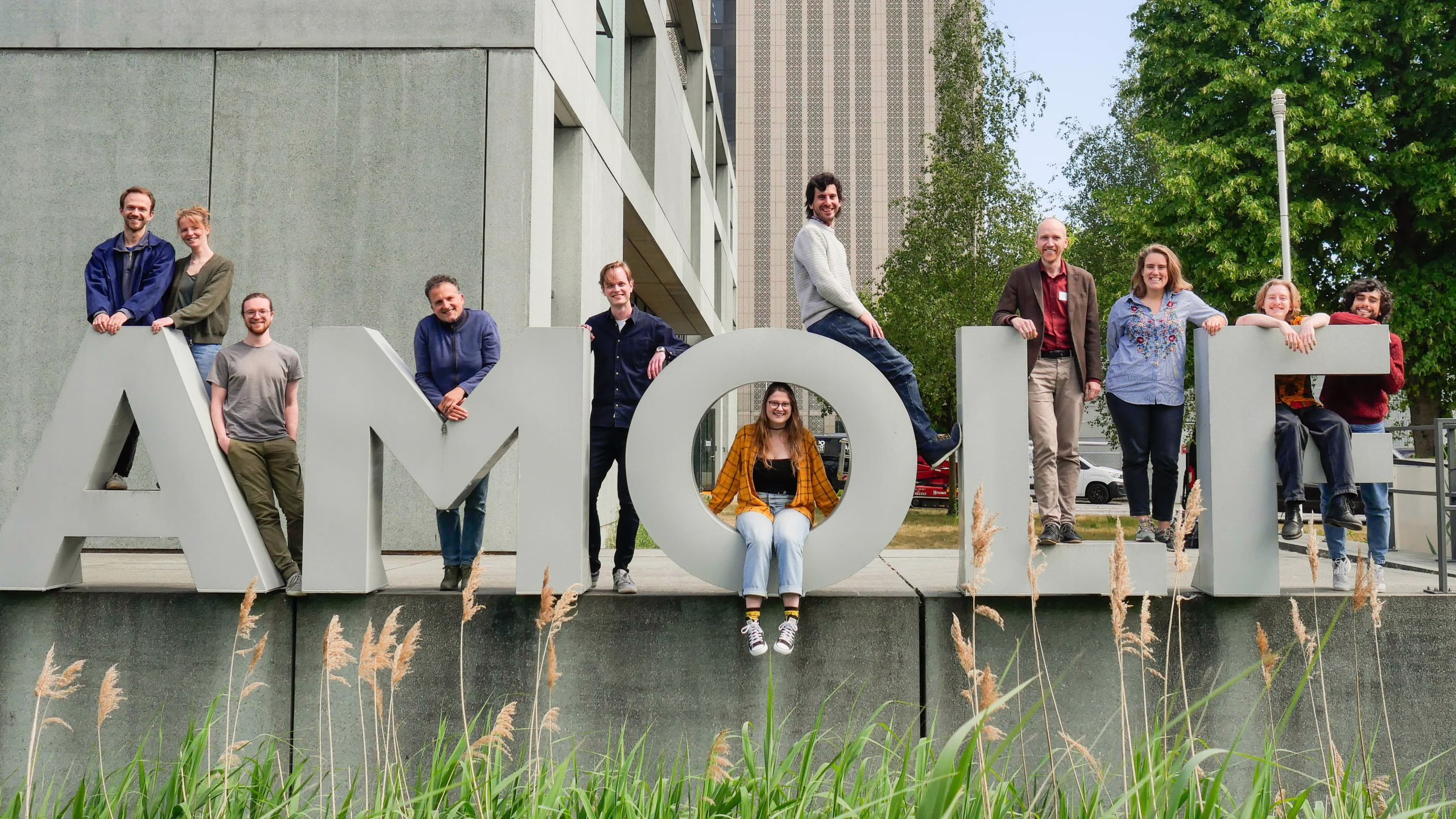 Groep mensen poseert bij grote letters AMOLI met groene planten en moderne gebouwen op de achtergrond