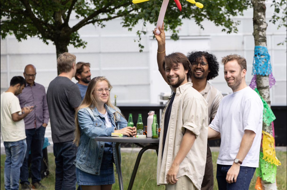 Colleagues standing and sitting around tables outdoors during the AMOLF BBQ, with three colleagues smiling at the camera in the foreground.